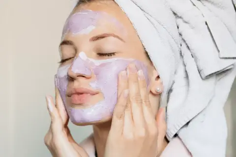 En kvinna med handduk i håret applicerar en lila ansiktsmask. Hon njuter av att ånga ansiktet för en avkopplande stund.